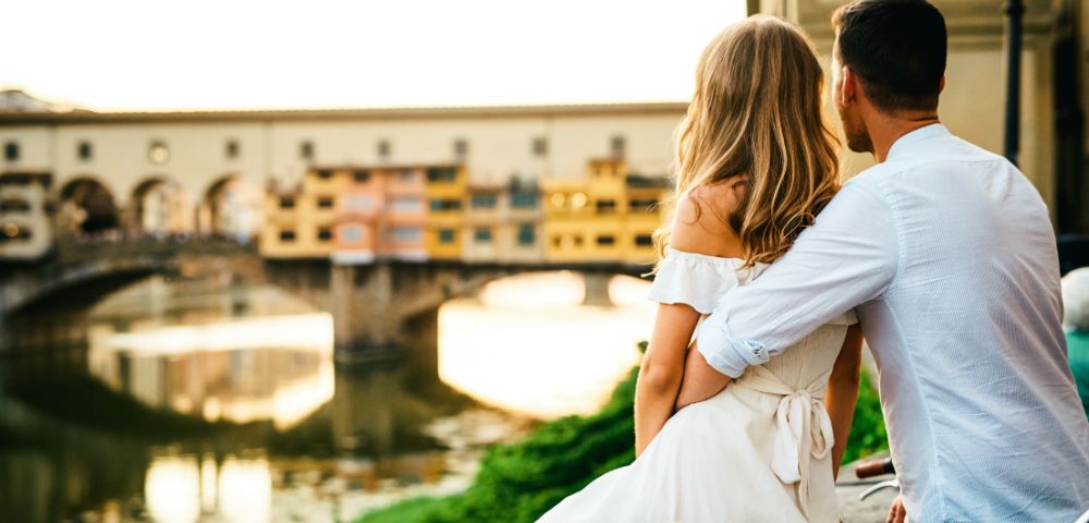 A couple sits closely, overlooking a river and historic bridge at sunset. They exude a romantic and peaceful vibe. A straw hat rests beside them.