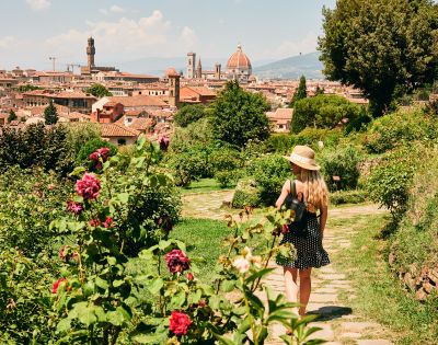 A woman in a polka-dot dress and sun hat walks through a lush garden filled with vibrant flowers, overlooking the picturesque cityscape of Florence, Italy.