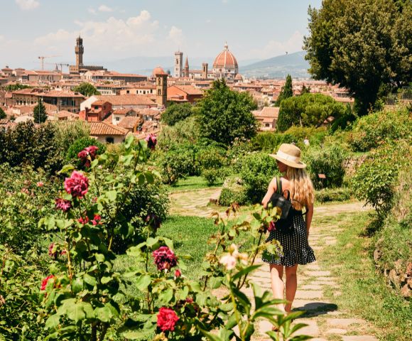 A woman in a polka-dot dress and sun hat walks through a lush garden filled with vibrant flowers, overlooking the picturesque cityscape of Florence, Italy.