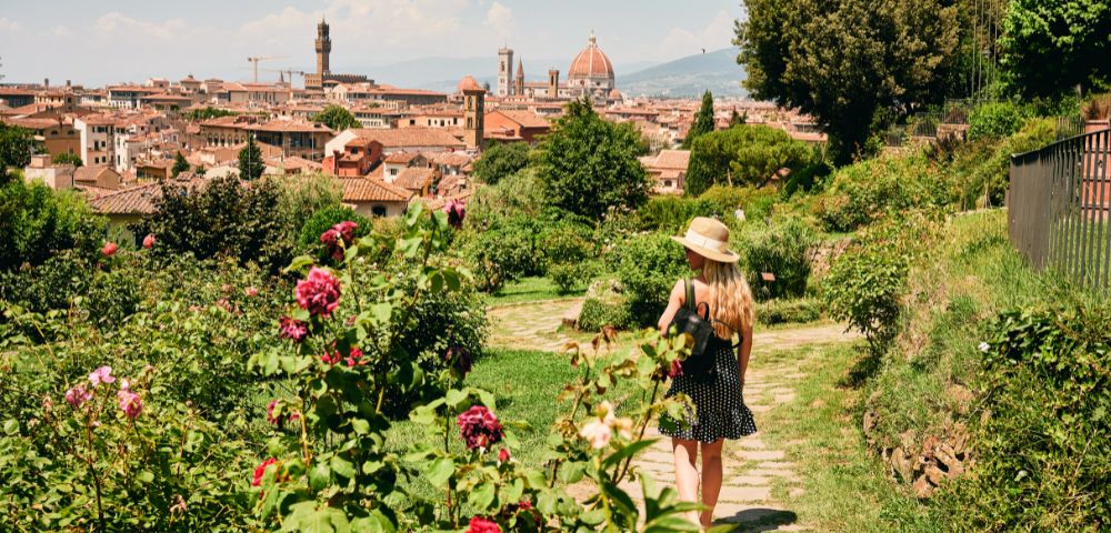 A woman in a polka-dot dress and sun hat walks through a lush garden filled with vibrant flowers, overlooking the picturesque cityscape of Florence, Italy.