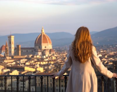 A woman with long hair, wearing a beige coat, looks over a Florence cityscape at sunset. The Duomo and distant hills are visible under a soft golden sky.