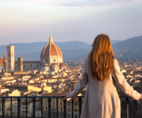 A woman with long hair, wearing a beige coat, looks over a Florence cityscape at sunset. The Duomo and distant hills are visible under a soft golden sky.