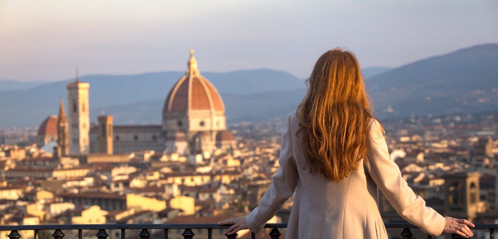 A woman with long hair, wearing a beige coat, looks over a Florence cityscape at sunset. The Duomo and distant hills are visible under a soft golden sky.