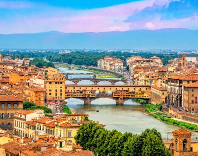 A scenic view of Florence, Italy, features the Ponte Vecchio bridge spanning the Arno River. Historic buildings and lush greenery frame the vibrant cityscape under a clear blue and pink sky.