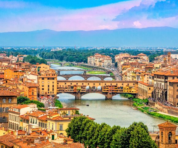 A scenic view of Florence, Italy, features the Ponte Vecchio bridge spanning the Arno River. Historic buildings and lush greenery frame the vibrant cityscape under a clear blue and pink sky.
