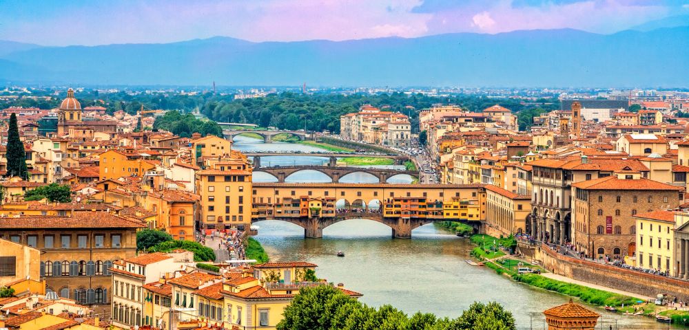 A scenic view of Florence, Italy, features the Ponte Vecchio bridge spanning the Arno River. Historic buildings and lush greenery frame the vibrant cityscape under a clear blue and pink sky.