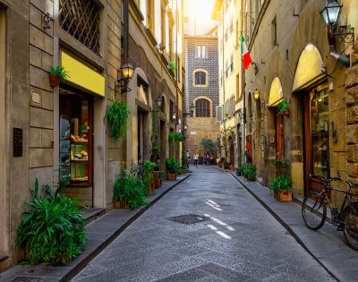 Charming narrow street lined with rustic buildings, shops, and greenery. Warm sunlight filters through, casting a cozy glow. Italian flag on right. Peaceful ambiance.