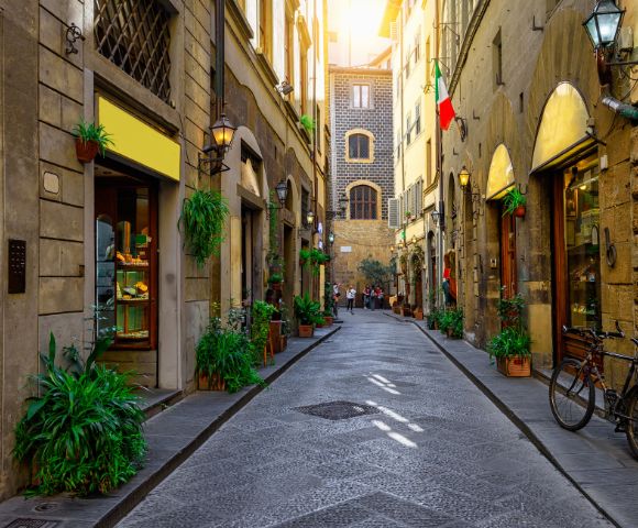 Charming narrow street lined with rustic buildings, shops, and greenery. Warm sunlight filters through, casting a cozy glow. Italian flag on right. Peaceful ambiance.