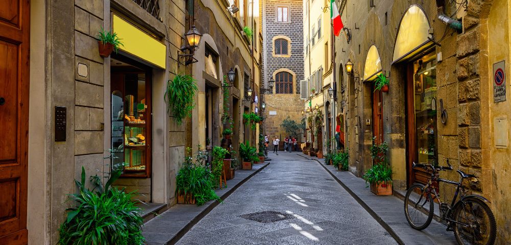 Charming narrow street lined with rustic buildings, shops, and greenery. Warm sunlight filters through, casting a cozy glow. Italian flag on right. Peaceful ambiance.