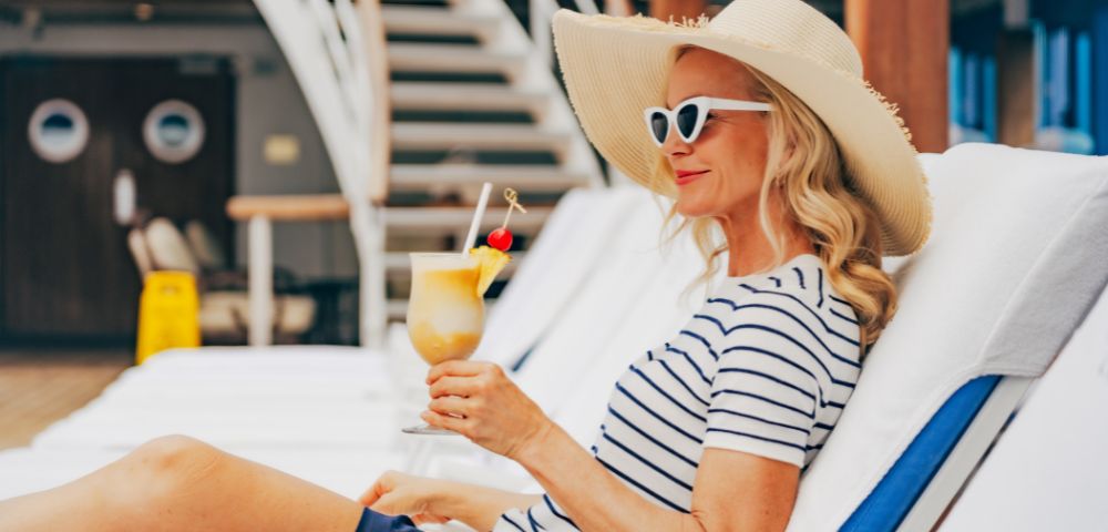 A woman in a striped shirt and sun hat lounges on a deck chair, holding a tropical drink. She exudes relaxation and enjoys a sunny, carefree setting.
