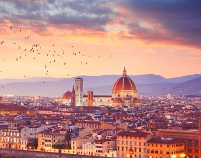 Sunset view of Florence with the Duomo's red dome, city rooftops, and flying birds. The sky glows with warm hues, creating a serene ambiance.