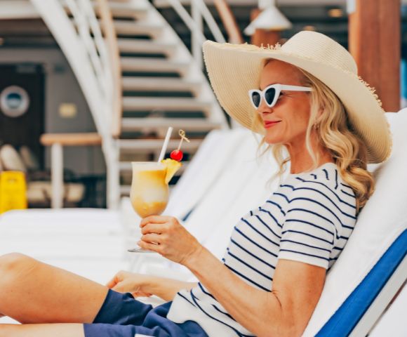 A woman in a striped shirt and sun hat lounges on a deck chair, holding a tropical drink. She exudes relaxation and enjoys a sunny, carefree setting.