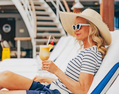 A woman in a striped shirt and sun hat lounges on a deck chair, holding a tropical drink. She exudes relaxation and enjoys a sunny, carefree setting.