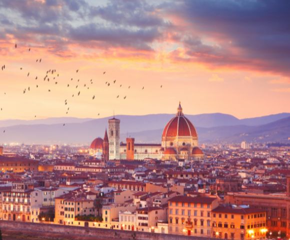 Sunset view of Florence with the Duomo's red dome, city rooftops, and flying birds. The sky glows with warm hues, creating a serene ambiance.