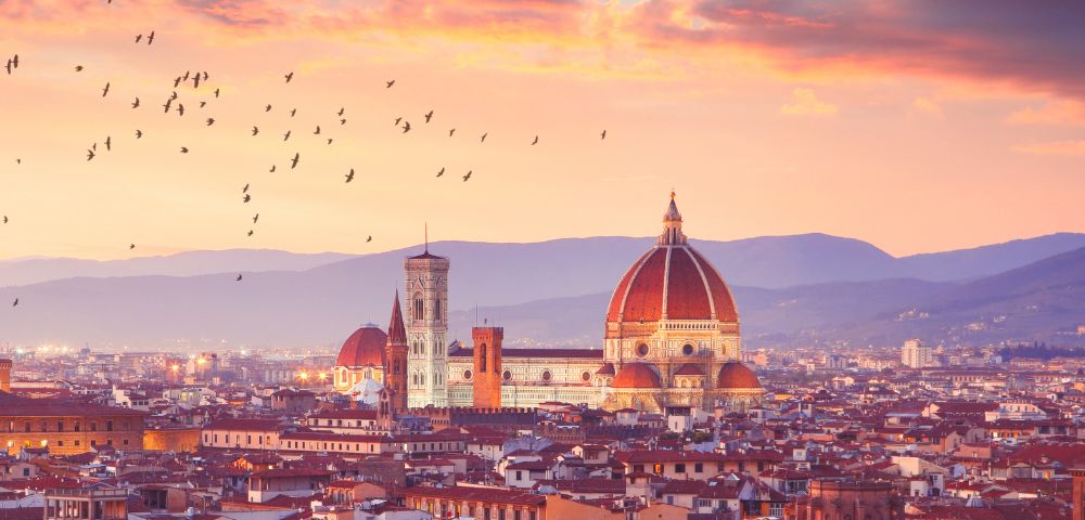 Sunset view of Florence with the Duomo's red dome, city rooftops, and flying birds. The sky glows with warm hues, creating a serene ambiance.