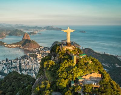 Aerial view of Rio de Janeiro with the Christ the Redeemer statue on a lush mountaintop. City buildings and Sugarloaf Mountain are visible against the ocean backdrop.