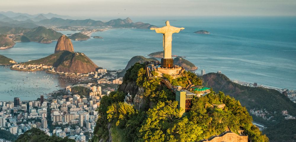 Aerial view of Rio de Janeiro with the Christ the Redeemer statue on a lush mountaintop. City buildings and Sugarloaf Mountain are visible against the ocean backdrop.