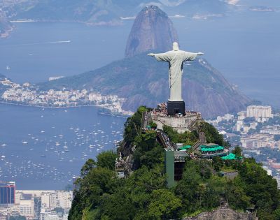 Aerial view of the Christ the Redeemer statue in Rio de Janeiro, Brazil, atop a green mountain. Cityscape and Sugarloaf Mountain are visible in the background.
