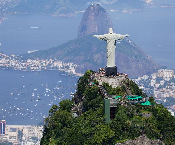 Aerial view of the Christ the Redeemer statue in Rio de Janeiro, Brazil, atop a green mountain. Cityscape and Sugarloaf Mountain are visible in the background.