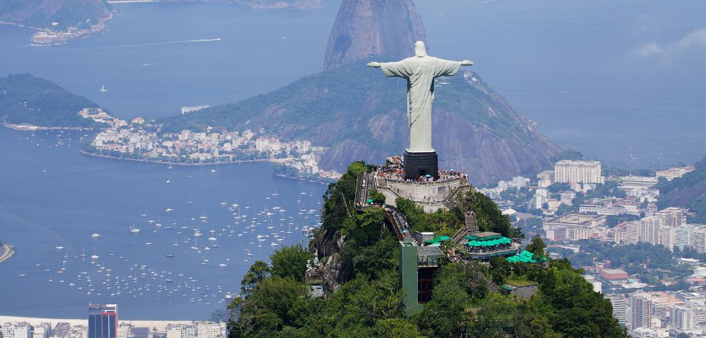 Aerial view of the Christ the Redeemer statue in Rio de Janeiro, Brazil, atop a green mountain. Cityscape and Sugarloaf Mountain are visible in the background.