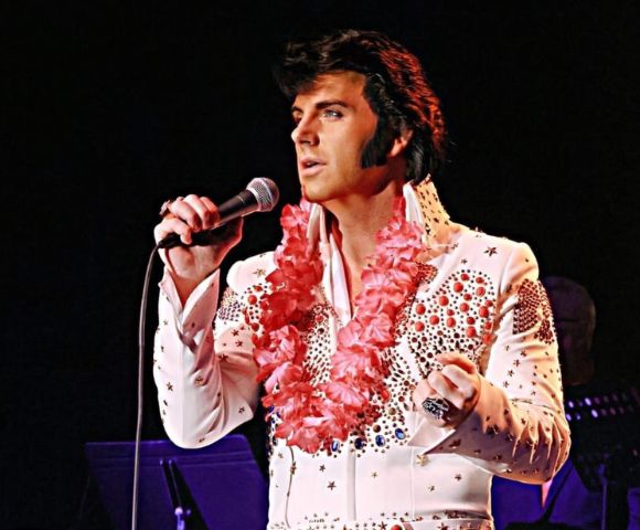 A performer in a sparkling white jumpsuit adorned with rhinestones and floral leis sings into a microphone on stage, with musicians behind.