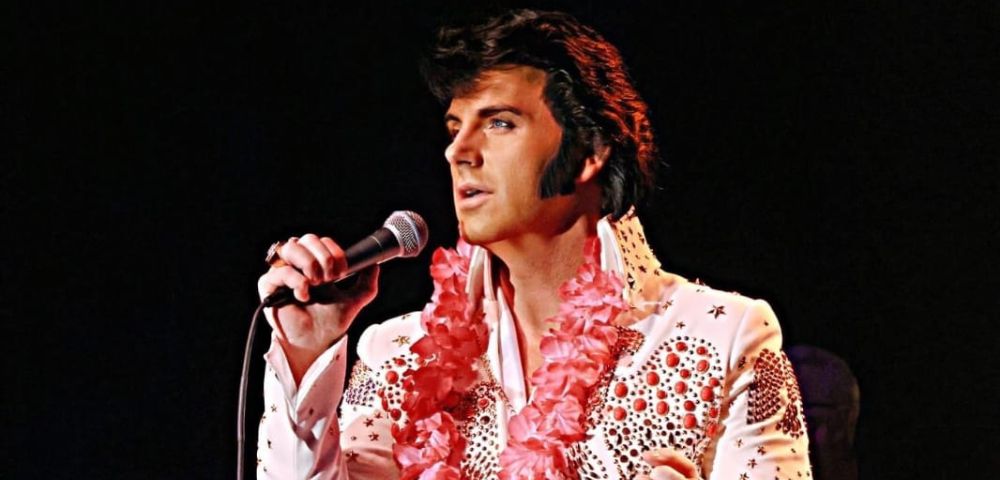 A performer in a sparkling white jumpsuit adorned with rhinestones and floral leis sings into a microphone on stage, with musicians behind.