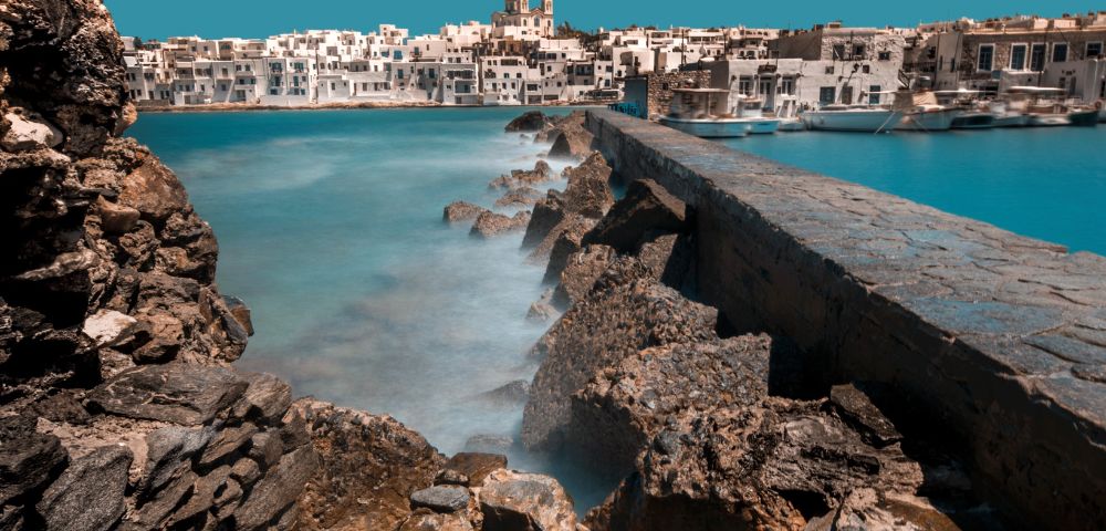 Scenic view of a coastal village with white buildings against a clear blue sky. A stone path runs along rugged rocks beside calm, turquoise water.