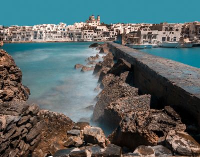 Scenic view of a coastal village with white buildings against a clear blue sky. A stone path runs along rugged rocks beside calm, turquoise water.