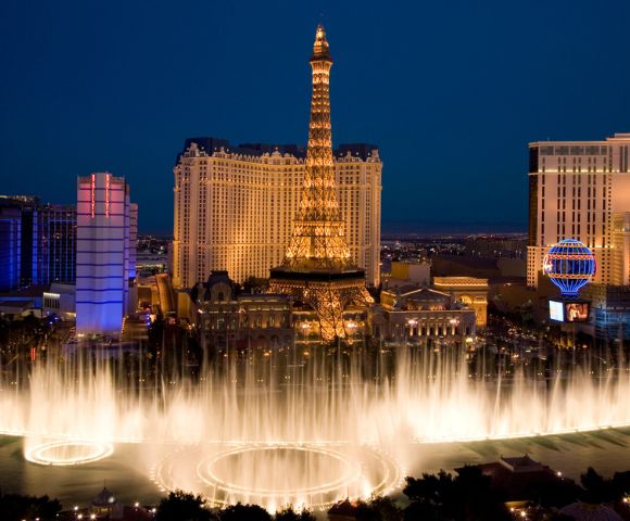 Night view of Las Vegas with illuminated fountains in the foreground and a replica Eiffel Tower in the background, creating an energetic, vibrant scene.
