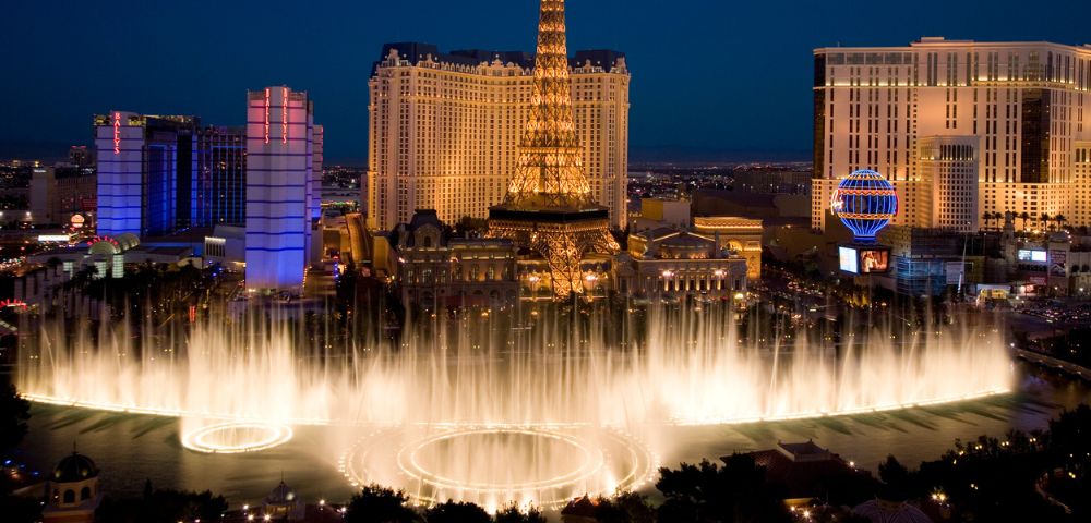 Night view of Las Vegas with illuminated fountains in the foreground and a replica Eiffel Tower in the background, creating an energetic, vibrant scene.