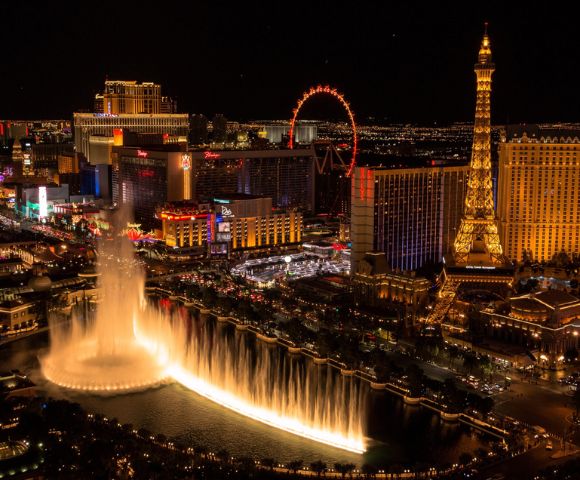 Night view of Las Vegas Strip featuring illuminated fountain, Eiffel Tower replica, bright neon lights, and lively, bustling atmosphere.
