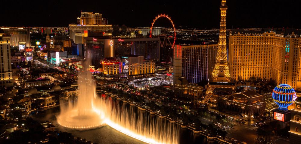 Night view of Las Vegas Strip featuring illuminated fountain, Eiffel Tower replica, bright neon lights, and lively, bustling atmosphere.