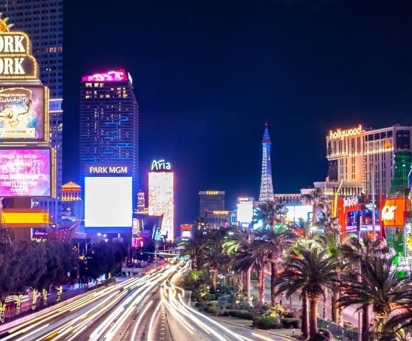 Bright Las Vegas Strip at night with illuminated signs for New York-New York and Park MGM. Vibrant lights and palm trees line the bustling street.
