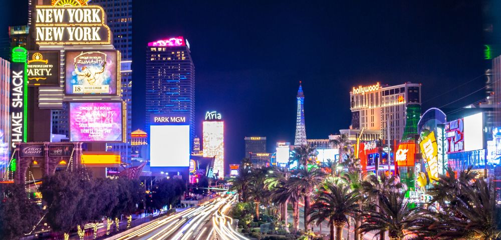 Bright Las Vegas Strip at night with illuminated signs for New York-New York and Park MGM. Vibrant lights and palm trees line the bustling street.