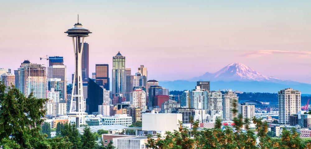 Skyline of Seattle at dusk with the prominent Space Needle, modern buildings, and Mt. Rainier in the background under a pastel pink and blue sky.