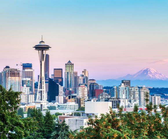 Skyline of Seattle at dusk with the prominent Space Needle, modern buildings, and Mt. Rainier in the background under a pastel pink and blue sky.