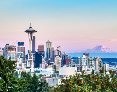 Skyline of Seattle at dusk with the prominent Space Needle, modern buildings, and Mt. Rainier in the background under a pastel pink and blue sky.