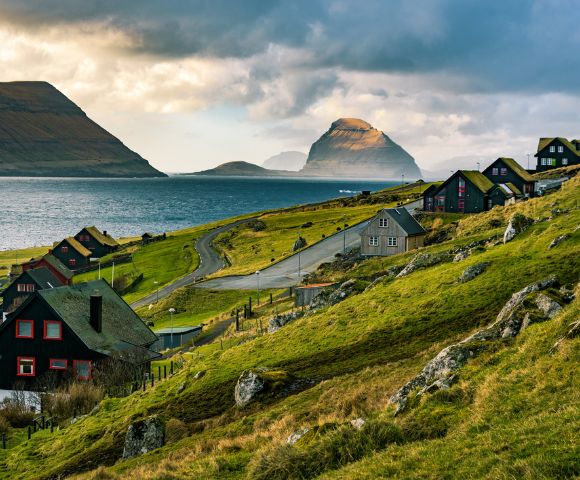 Scenic view of lush green hills with quaint houses by a winding road, leading to a tranquil sea. Majestic mountains rise under a dramatic cloudy sky.