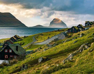 Scenic view of lush green hills with quaint houses by a winding road, leading to a tranquil sea. Majestic mountains rise under a dramatic cloudy sky.