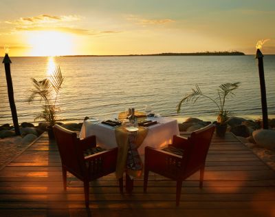 A romantic oceanfront dinner setup at sunset shows two chairs at a table with a white tablecloth on a wooden deck, surrounded by tiki torches and palm plants.