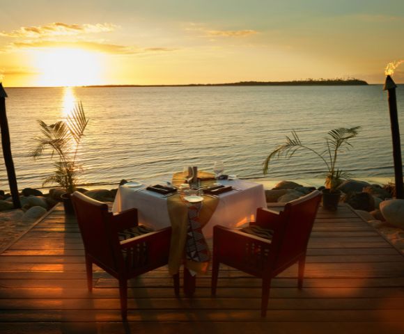 A romantic oceanfront dinner setup at sunset shows two chairs at a table with a white tablecloth on a wooden deck, surrounded by tiki torches and palm plants.