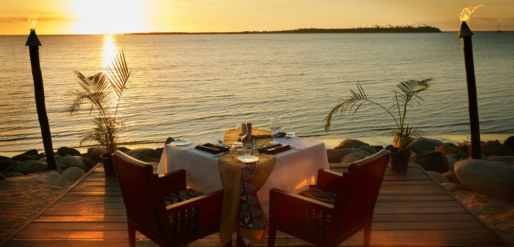 A romantic oceanfront dinner setup at sunset shows two chairs at a table with a white tablecloth on a wooden deck, surrounded by tiki torches and palm plants.