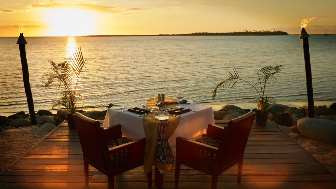A romantic oceanfront dinner setup at sunset shows two chairs at a table with a white tablecloth on a wooden deck, surrounded by tiki torches and palm plants.
