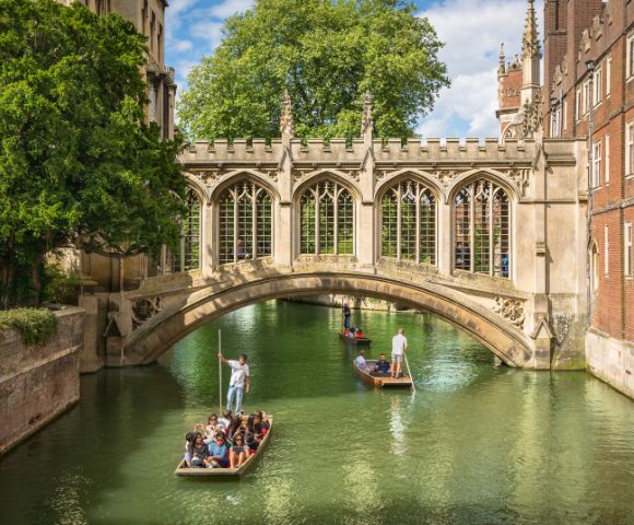 People enjoy a leisurely punt ride on a serene river under a historic stone bridge, surrounded by lush greenery and Gothic architecture on a sunny day.