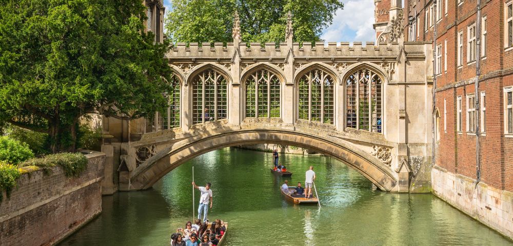 People enjoy a leisurely punt ride on a serene river under a historic stone bridge, surrounded by lush greenery and Gothic architecture on a sunny day.