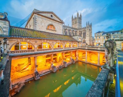 Historic Roman Baths in Bath, England, at dusk. Warm lights illuminate the arches and statues, with a cathedral visible in the background.