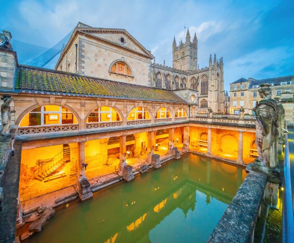 Historic Roman Baths in Bath, England, at dusk. Warm lights illuminate the arches and statues, with a cathedral visible in the background.