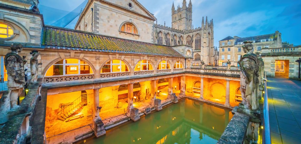 Historic Roman Baths in Bath, England, at dusk. Warm lights illuminate the arches and statues, with a cathedral visible in the background.