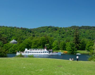 A serene landscape featuring a white boat cruising on a tranquil river, surrounded by lush green hills and trees. People sit on the grassy bank under a clear blue sky, evoking a peaceful, leisurely vibe.