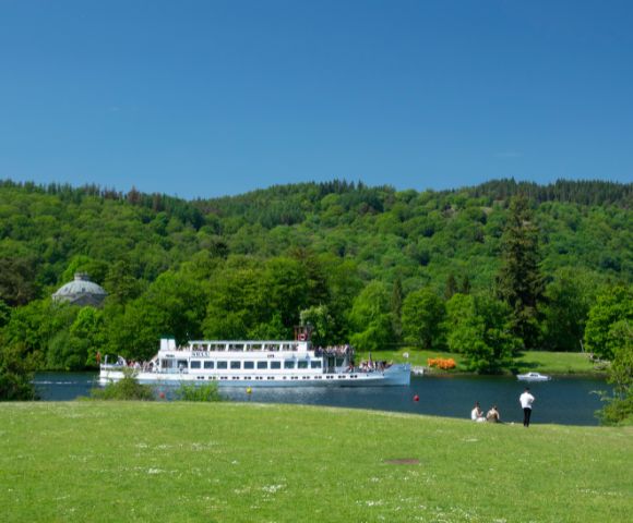 A serene landscape featuring a white boat cruising on a tranquil river, surrounded by lush green hills and trees. People sit on the grassy bank under a clear blue sky, evoking a peaceful, leisurely vibe.